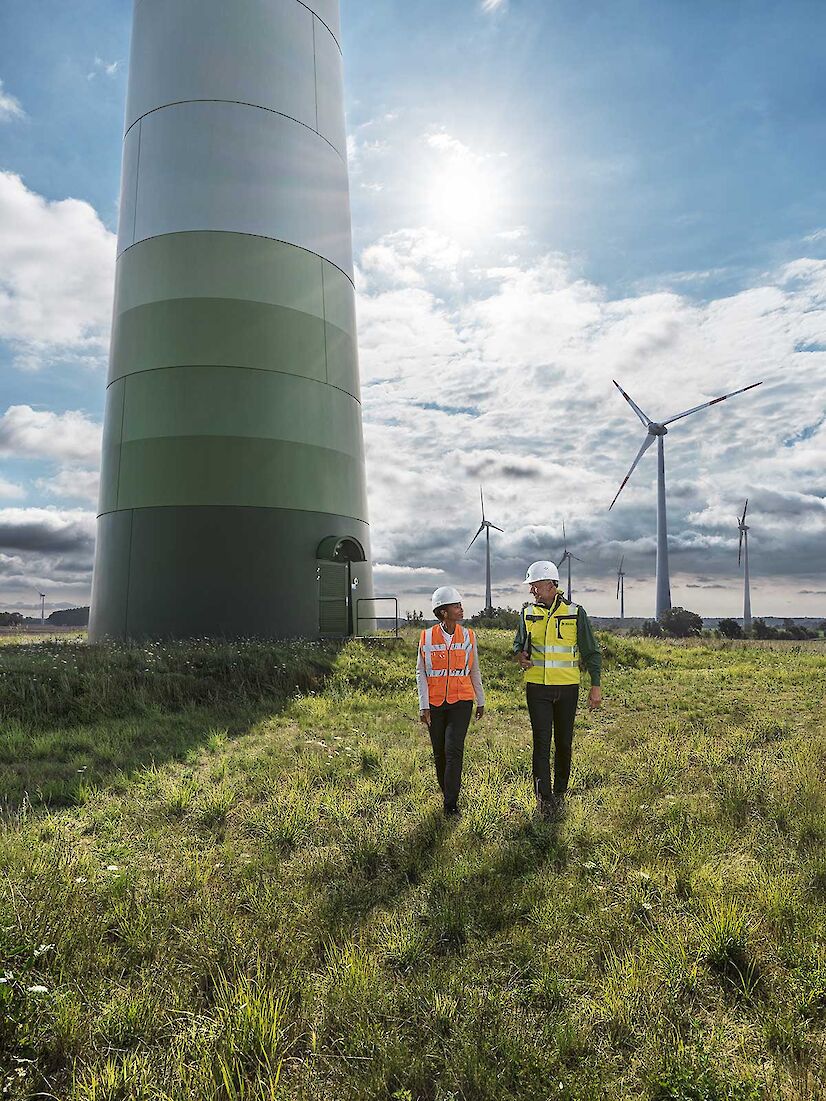 An engineer and a DEKRA engineer are walking across a meadow. Several wind turbines can be seen behind them.