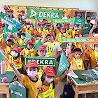 A classroom with lots of children wearing DEKRA children's caps and holding shields with DEKRA written on them.